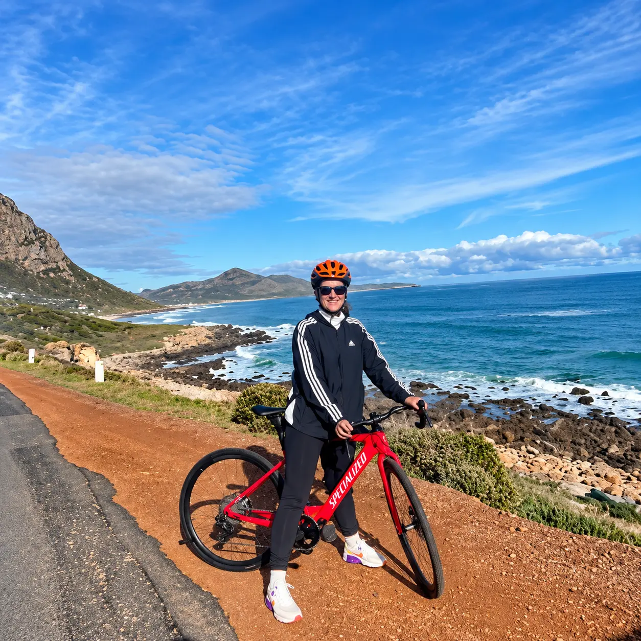 Cyclist with a red bicycle overlooking a rocky coastline and blue ocean on a scenic Cape Peninsula cycling tour.